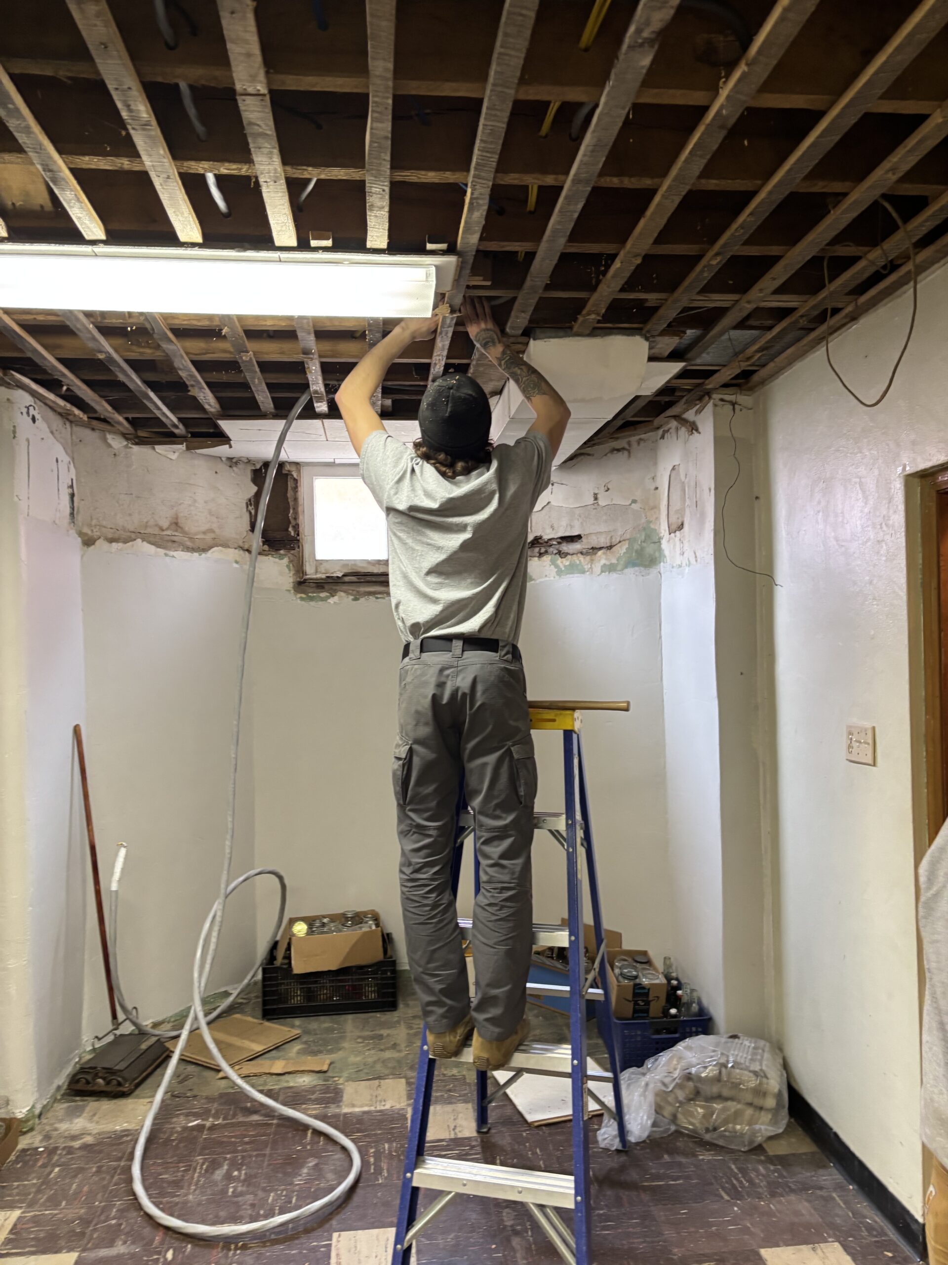 A person standing on a ladder installs or repairs electrical wiring on an exposed ceiling in a partially renovated room.