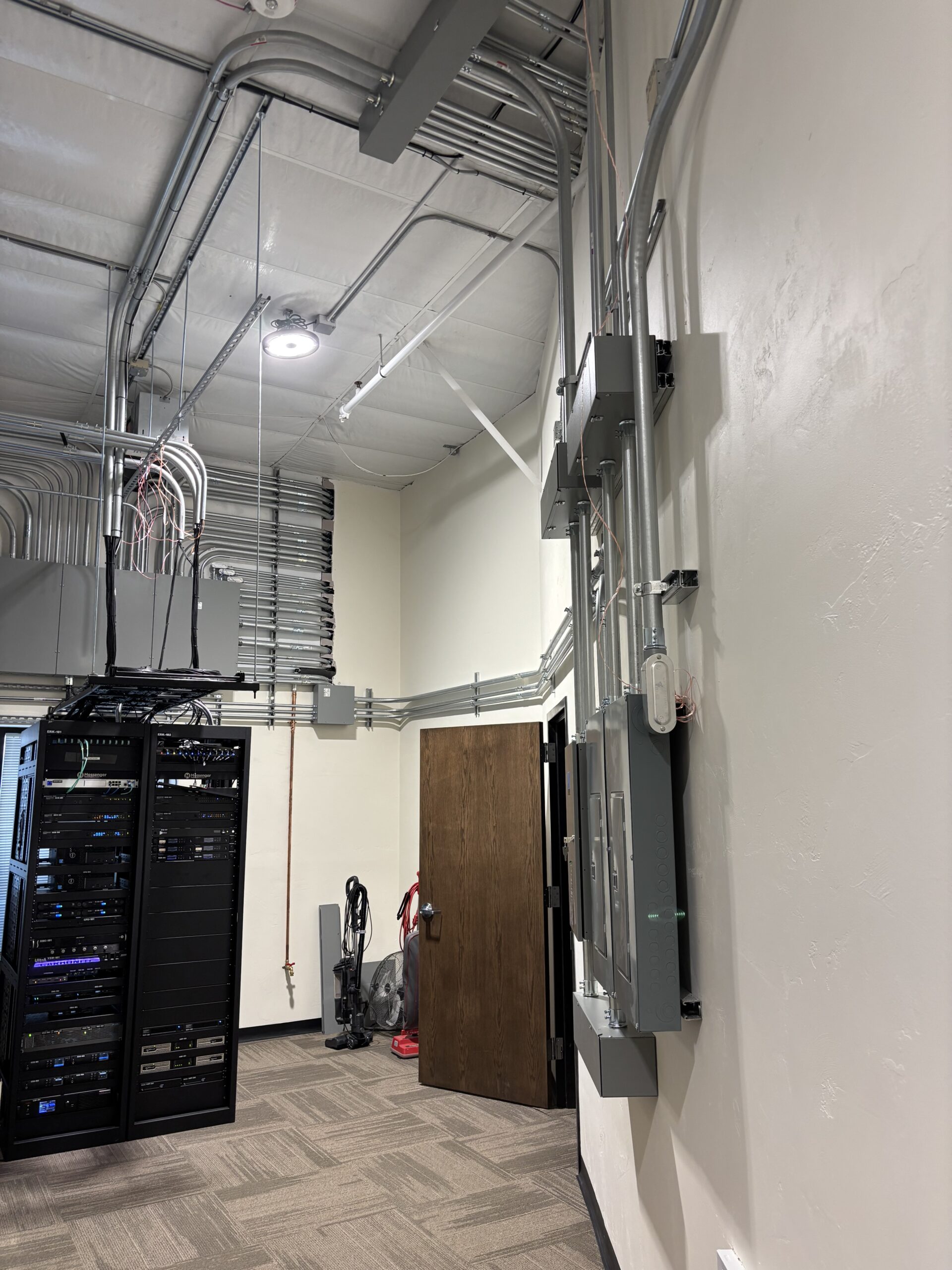 A server room with network racks, exposed cables and conduits on the walls and ceiling, an open door, and cleaning equipment in the corner.