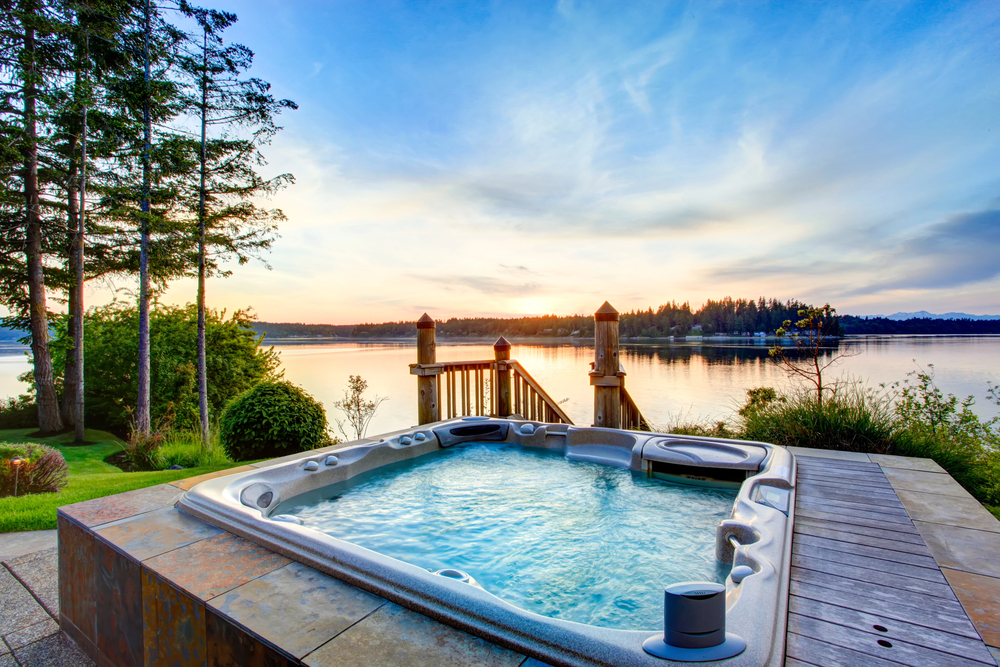 Outdoor hot tub on a patio overlooking a calm lake with trees and a wooden dock at sunset.