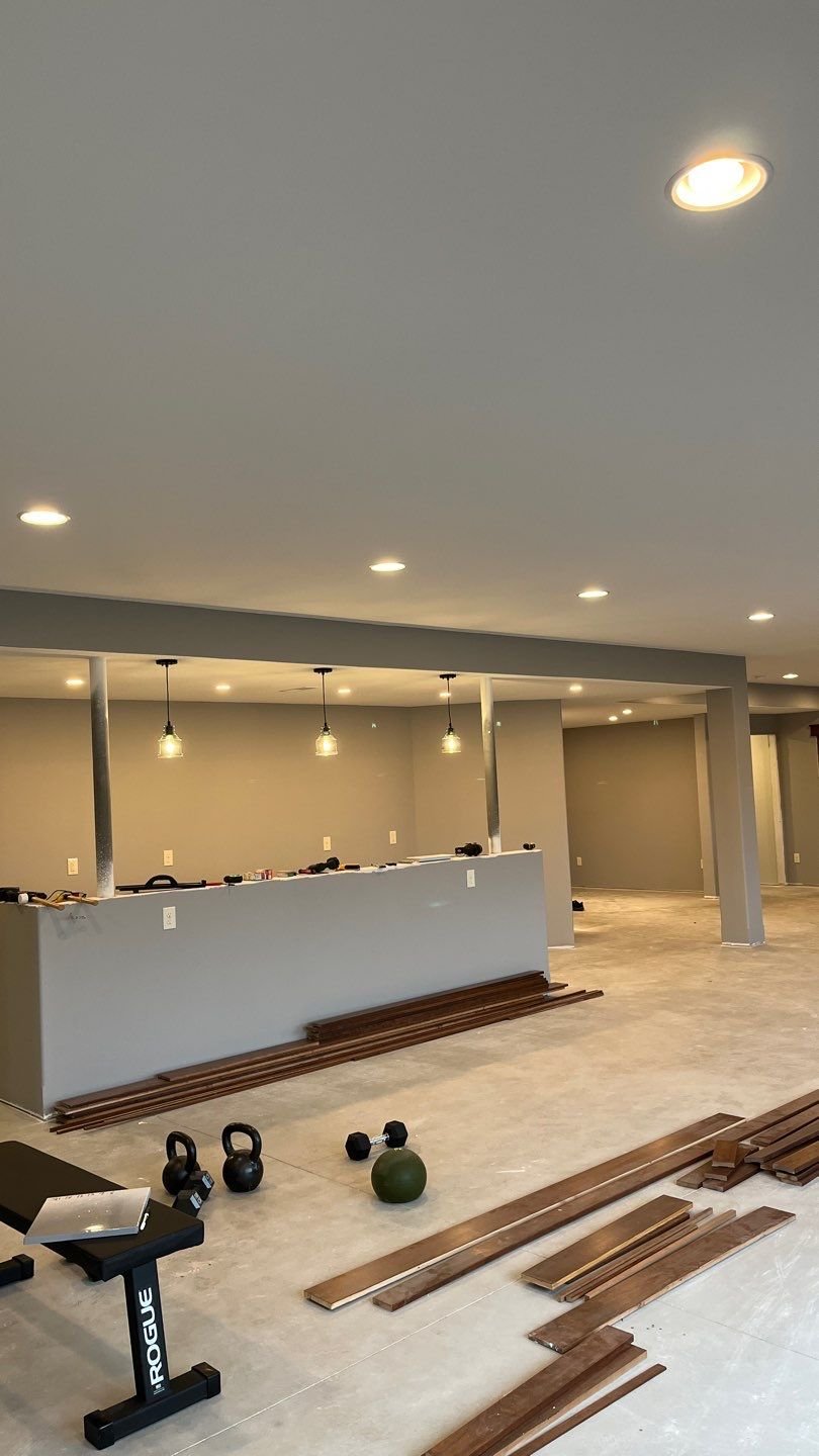 A partially finished basement with recessed lighting, wood flooring planks, kettlebells, a bench, and a bar area with hanging pendant lights.