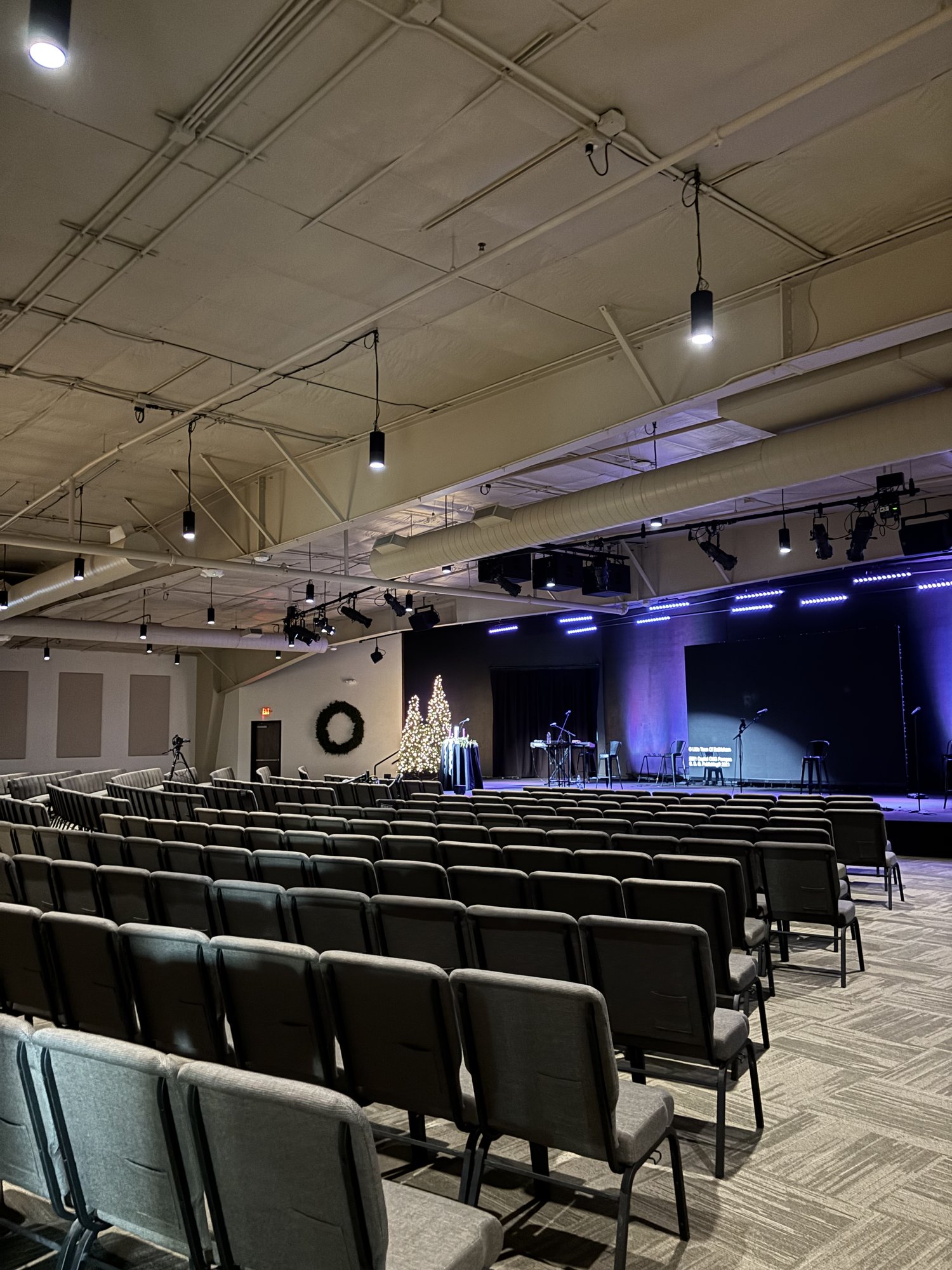Rows of empty chairs face a stage with instruments and music stands in a large, modern auditorium with overhead lights.