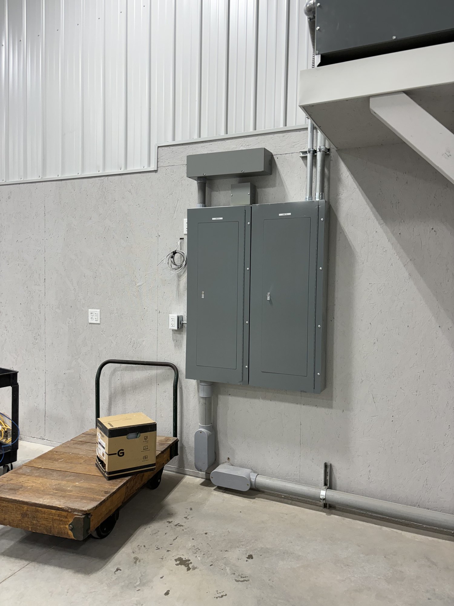 Two gray electrical panels are mounted on a light-colored wall in an industrial space, with conduit pipes connected above and below. A wooden cart with a cardboard box sits nearby.