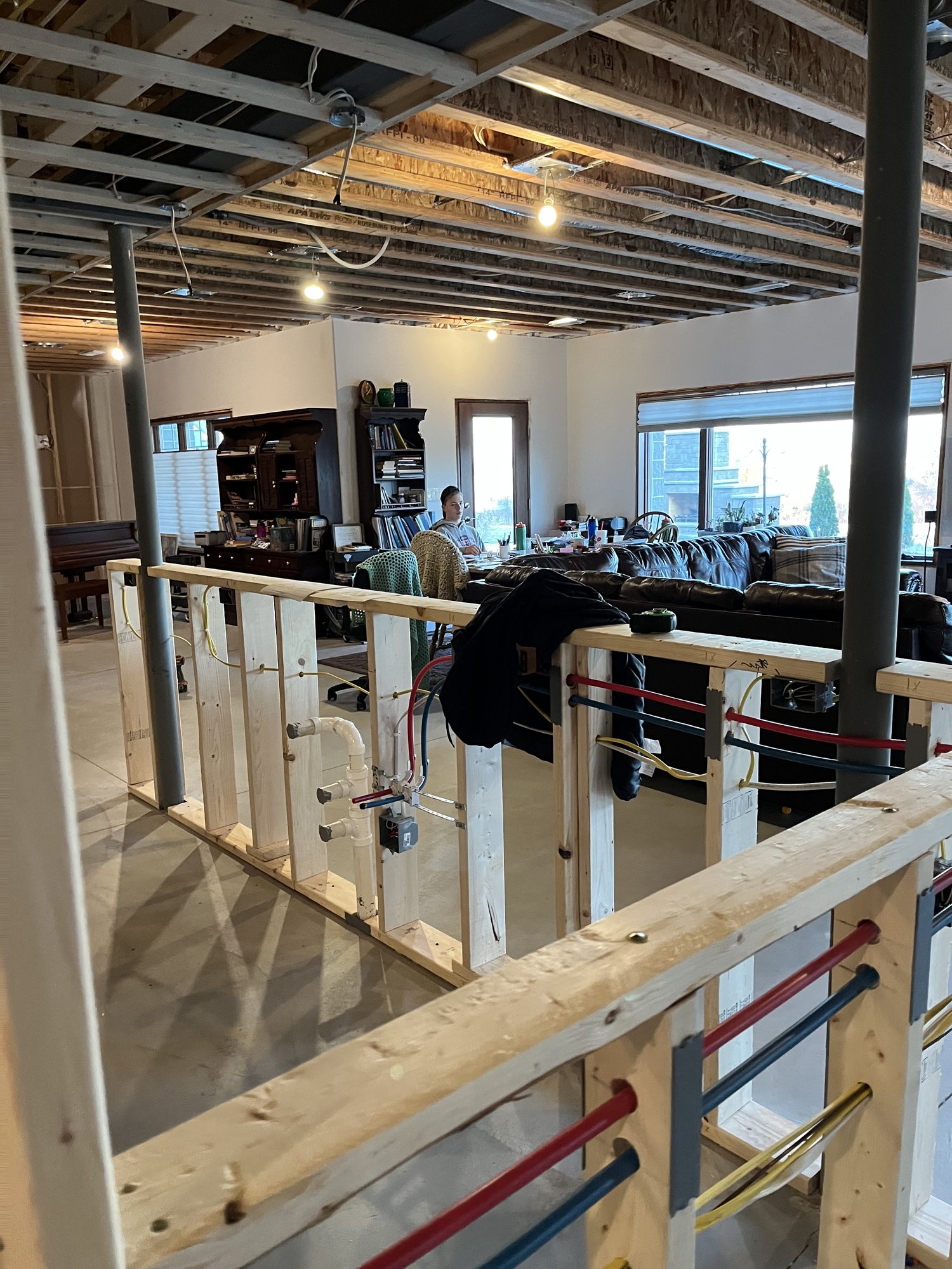 A person working on a laptop at a table in a partially finished room with exposed wooden framing, ceiling beams, and visible pipes.